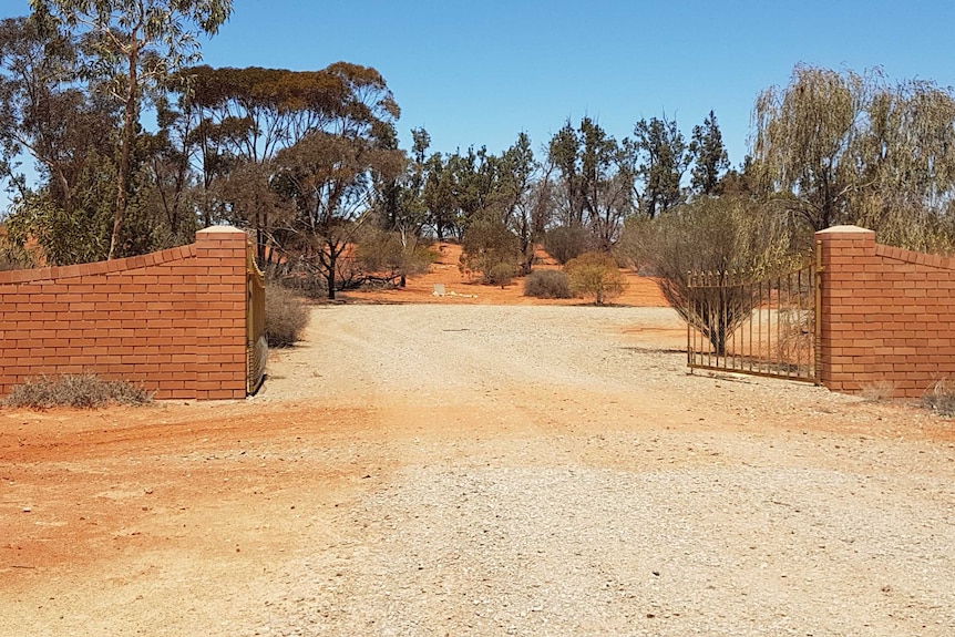 Brick walls hold gates opening to a red dirt cemetery, where only a few small plots can be seen in the middle of the image