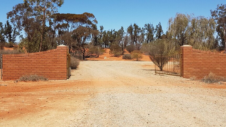 Brick walls hold gates opening to a red dirt cemetery, where only a few small plots can be seen in the middle of the image