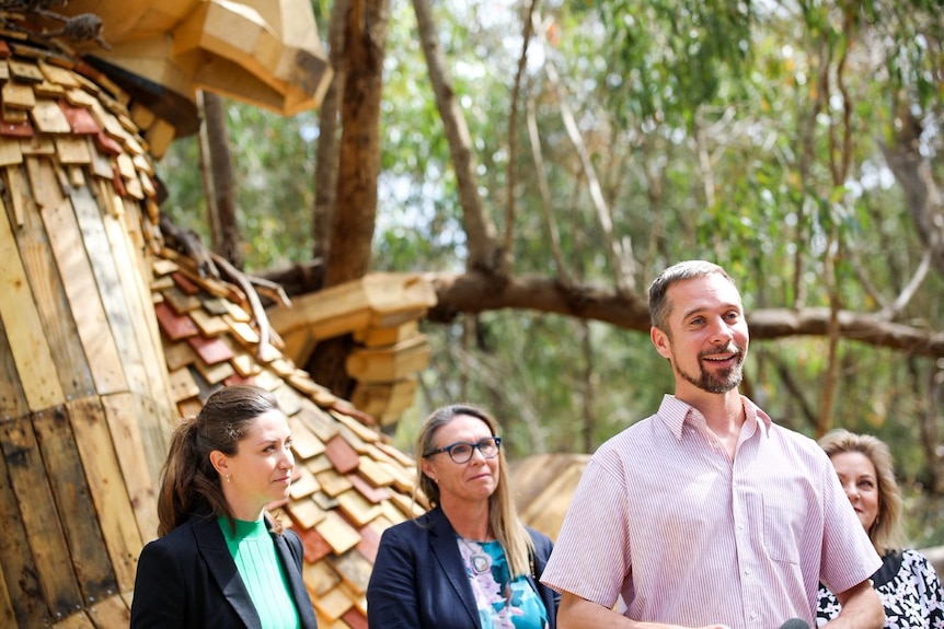 Thomas smiling and standing in front of a large wooden sculpture 