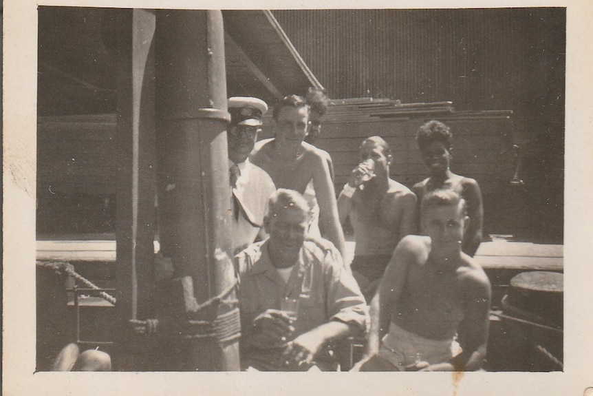 Five men, three of them shirtless, pose for a photo while having a drink on a ship. 