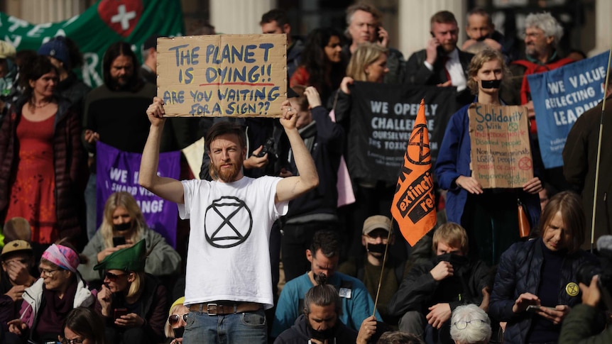 A large group of protestors hold up placards one in the centre reads "the planet is dying are you waiting or a sign"