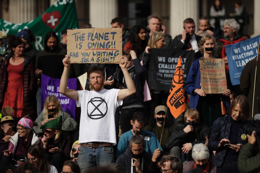 A large group of protestors hold up placards one in the centre reads "the planet is dying are you waiting or a sign"