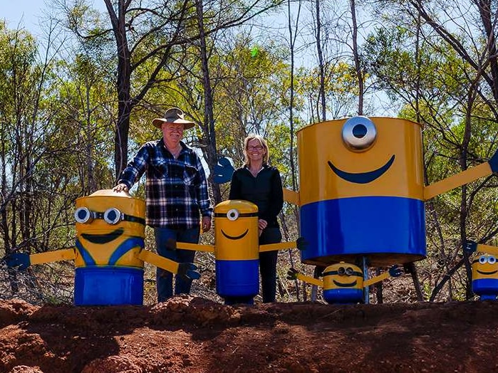 A couple stand in Australian bushland, red dirt in foreground, surrounded by home-made minions.