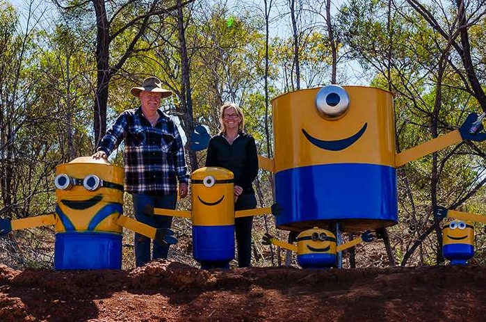 A couple stand in Australian bushland, red dirt in foreground, surrounded by home-made minions.