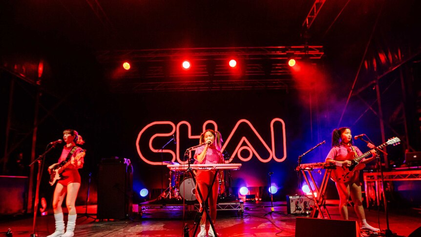 Three Japanese women singing into mics and playing bass guitar, keys and guitar on stage, 'Chai' in neon in the b/g