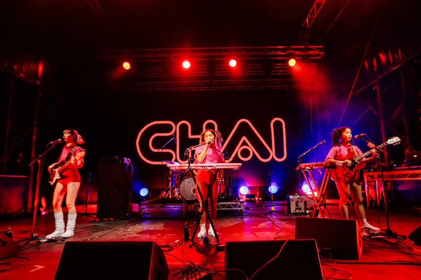 Three Japanese women singing into mics and playing bass guitar, keys and guitar on stage, 'Chai' in neon in the b/g