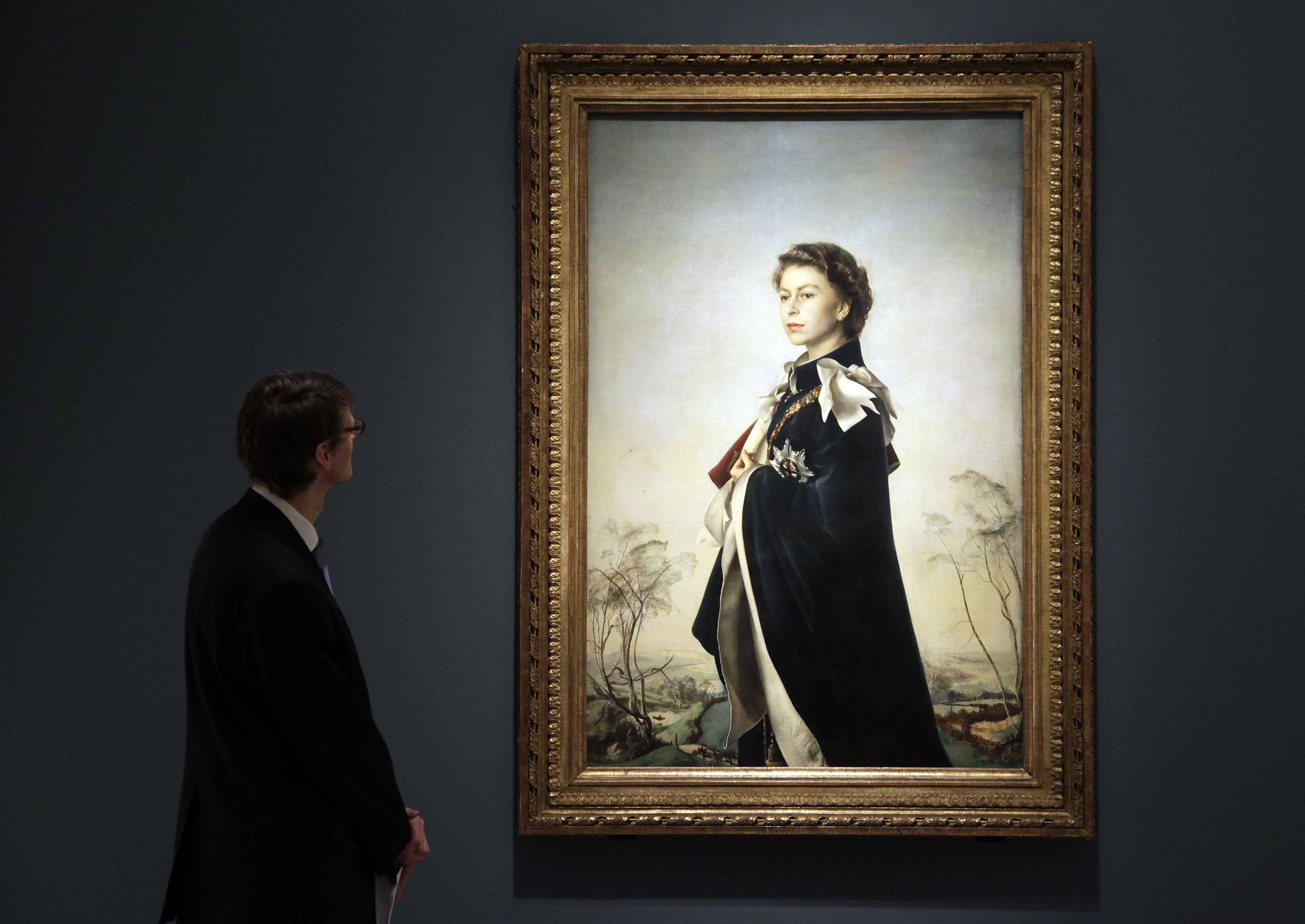 A man in a suit stands to the left of a lit-up, wall-mounted painting of a young, regal-looking Queen Elizabeth.