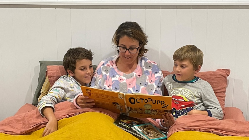 A woman sits between two young boys reading them a storybook.