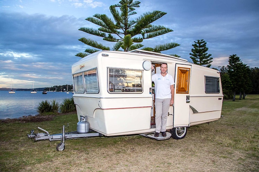 Justin Hales of Camplify with his caravan standing against a beachscape