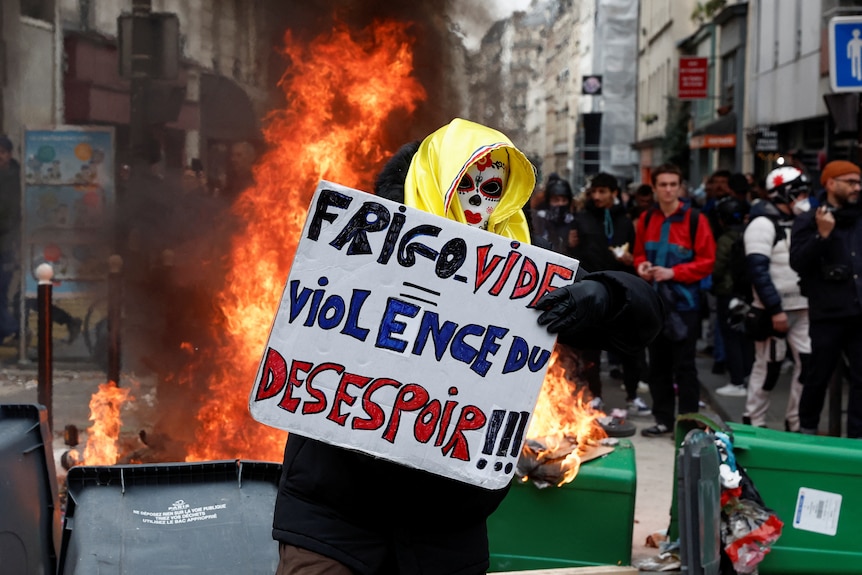 A person wearing a mask holdsup a sign while standing in front of a fire.