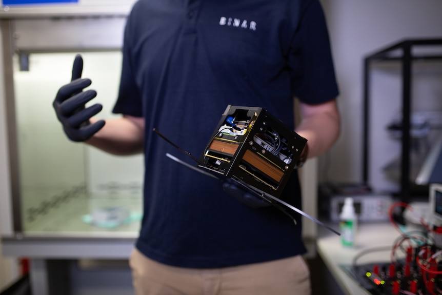 A standing in a scientific lab man holds a small black cube in his left hand.