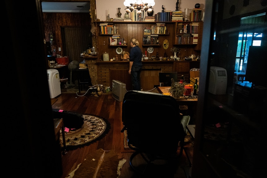 A woman walks through a living, lightly cluttered living room with wooden floorboards.