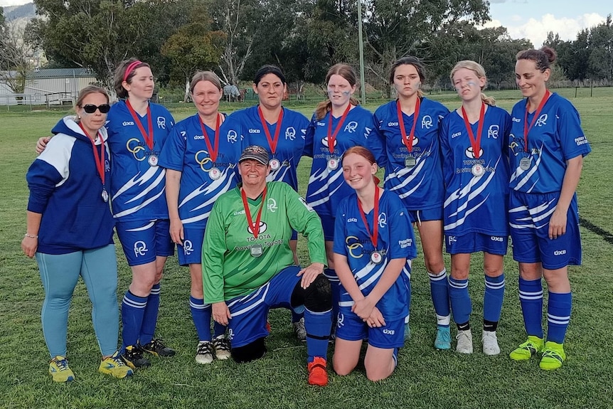 Short-haired woman wears cap,green jersey, in group photo with teammates wearing blue jerseys, medals around neck, green field.