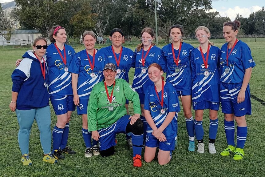 Short-haired woman wears cap,green jersey, in group photo with teammates wearing blue jerseys, medals around neck, green field.