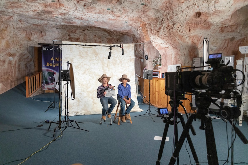 Two children wearing cowboy hats sitting on stools in front of TV equipment in a cave.