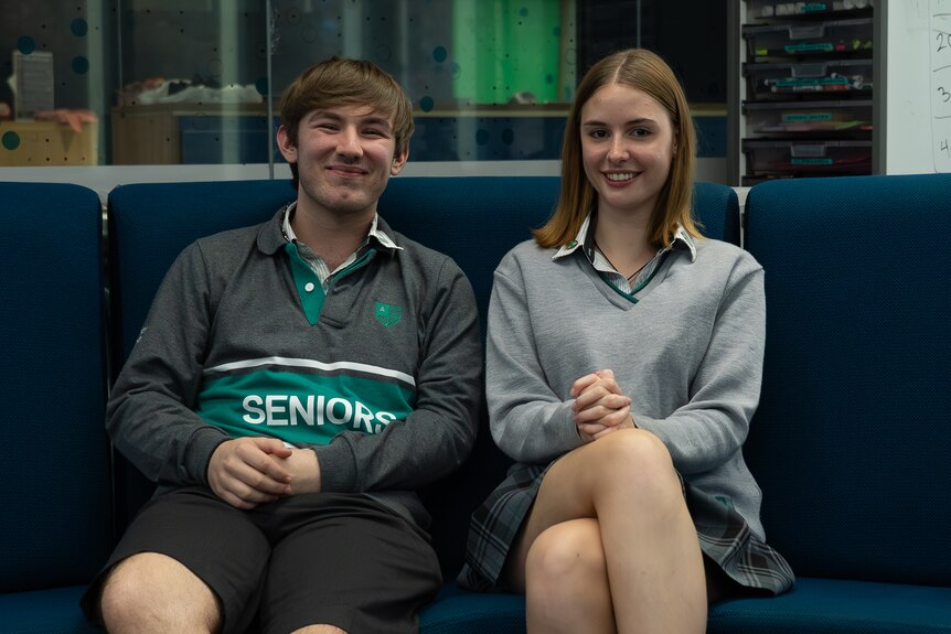 A teenage boy and girl, both smiling, sit next to each other on a couch.