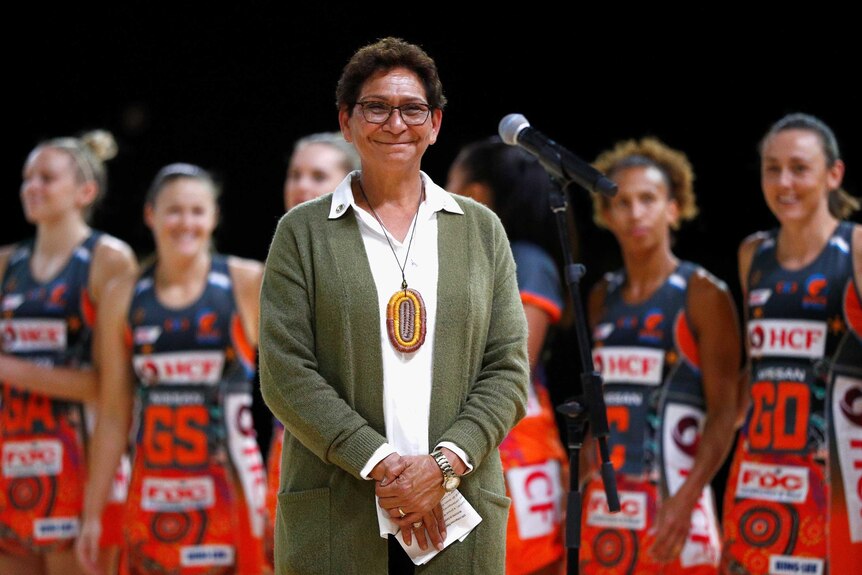 A smiling Indigenous lady stands near a microphone as a team of netballers stand behind her.