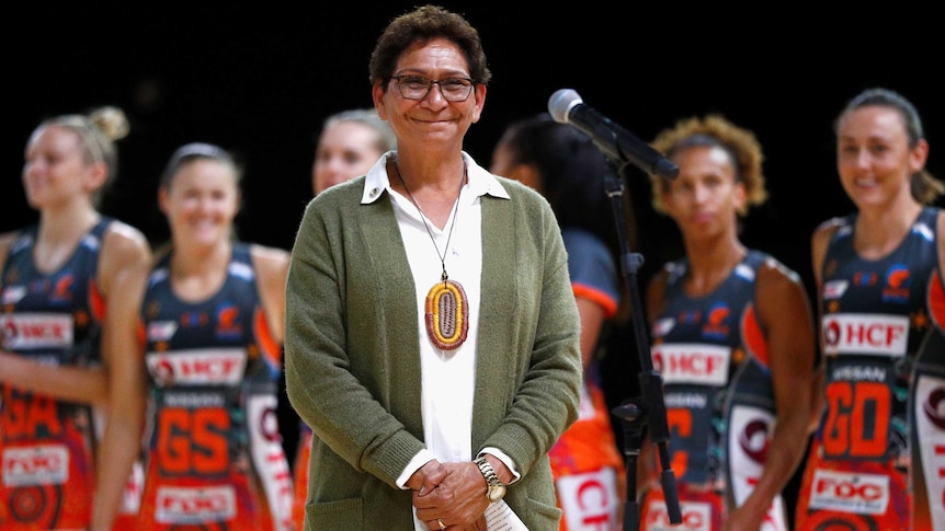 A smiling Indigenous lady stands near a microphone as a team of netballers stand behind her.