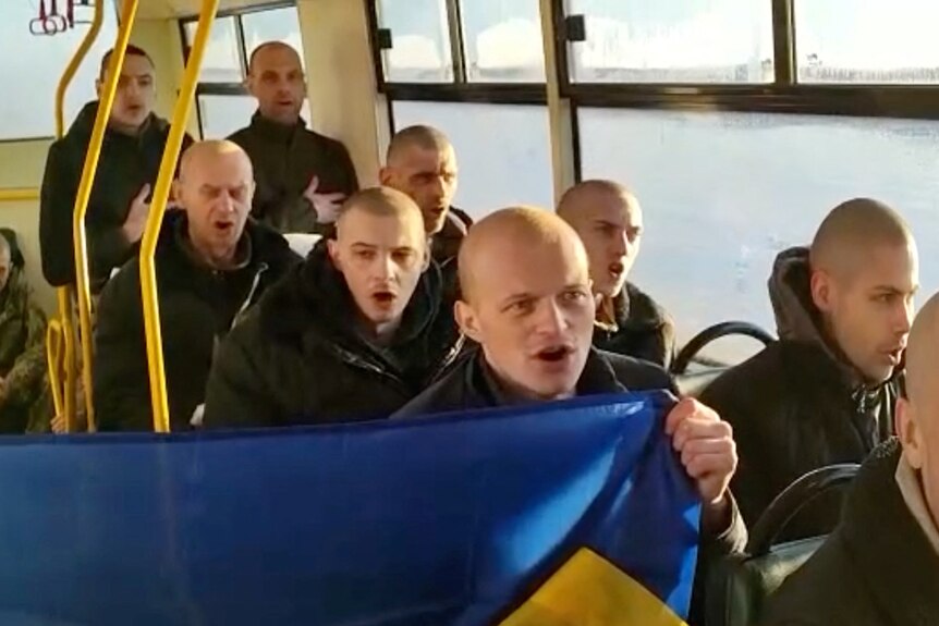 Bald men sing while seated on a bus, holding a Ukrainian flag.