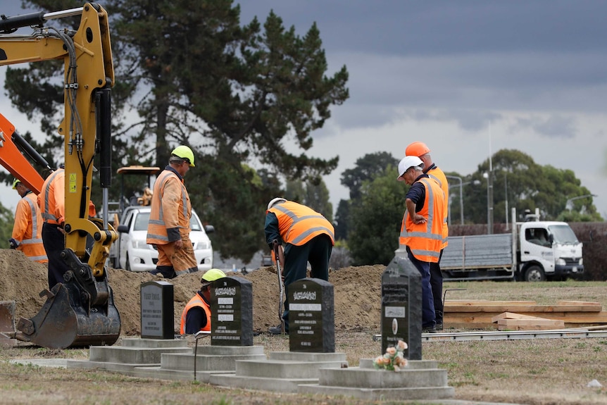 Workers wearing high-visibility clothing use shovels and a backhoe to dig graves at a cemetery.