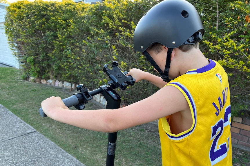 A young boy, wearing a helmet stands on an e-scooter