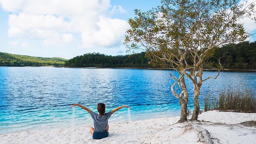 Woman sits on white sand beside very blue lake with sand pouring from her hands