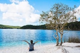 Woman sits on white sand beside very blue lake with sand pouring from her hands