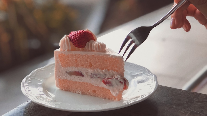 A hand holding a fork hovers over a pink sponge cake, filled with cream, with icing and a strawberry on top