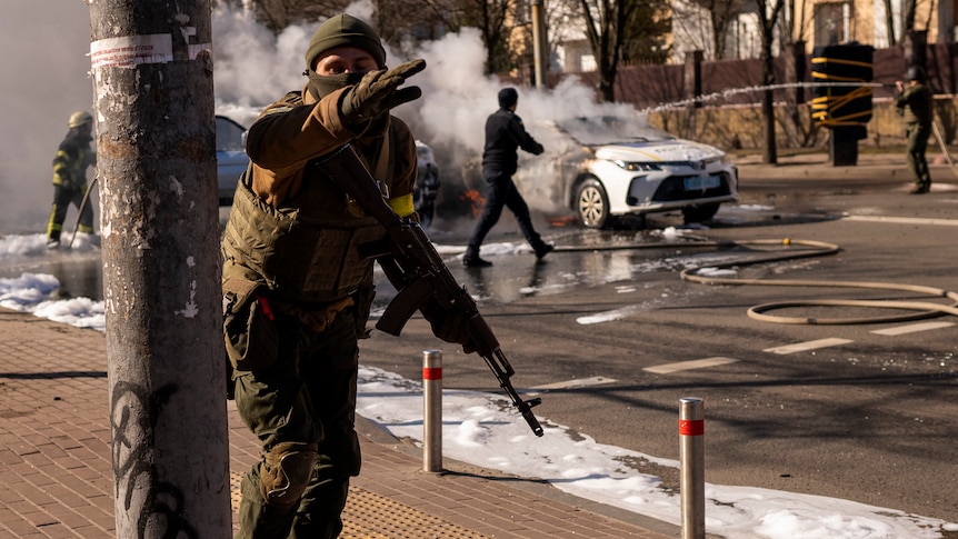 An armed Ukrainian soldier points forward with his left arm as he and other troops move past burning cars on a asphalt road.
