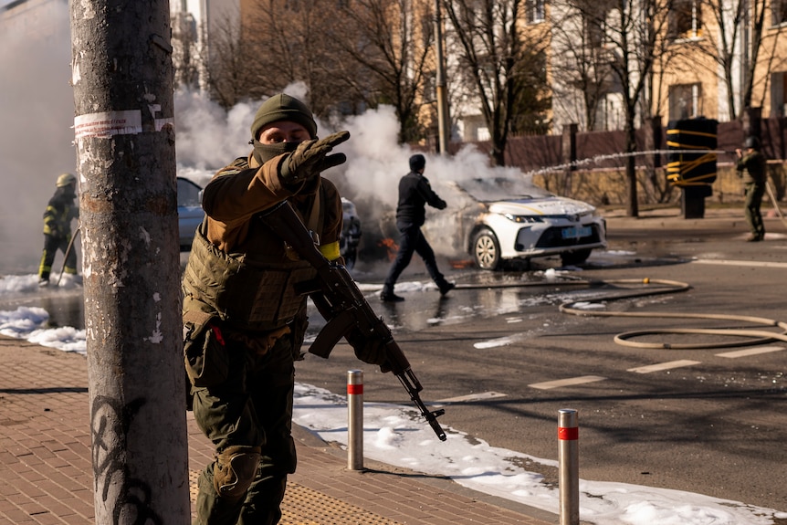 An armed Ukrainian soldier points forward with his left arm as he and other troops move past burning cars on a asphalt road.