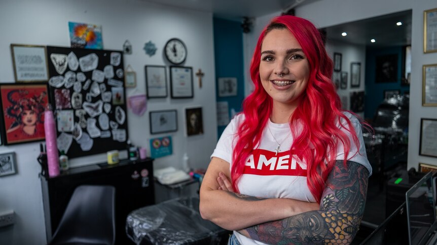 Woman with a large tattoo along her right arm and bright red hair, standing in a tattoo parlour with her arms folded.