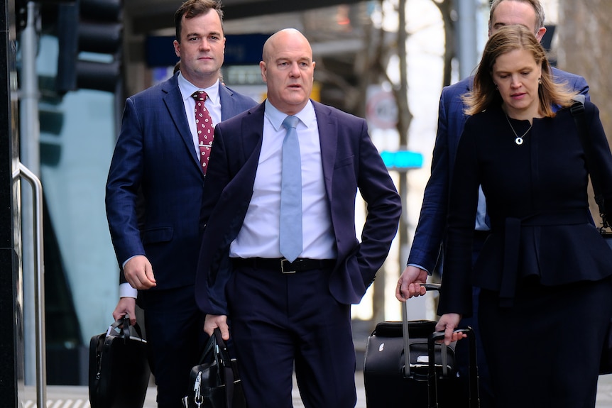  A bald man wearing a blue suit, light blue shirt and blue tie holds a briefcase, surrounded by two men and a woman.