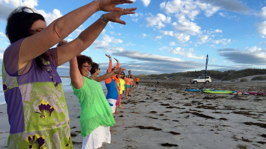 A group of people dressed in fluoro colours stand on a beach in a line doing the mexican wave.