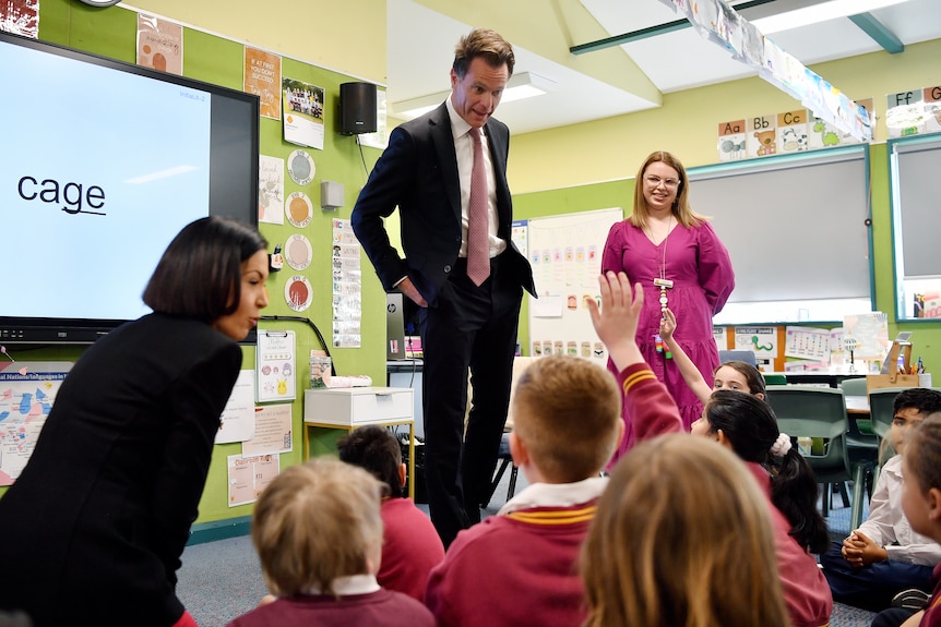 El primer ministro de Nueva Gales del Sur, Chris Minns, dentro de un aula hablando con los estudiantes que están sentados en el suelo en la escuela pública de Riverstone.