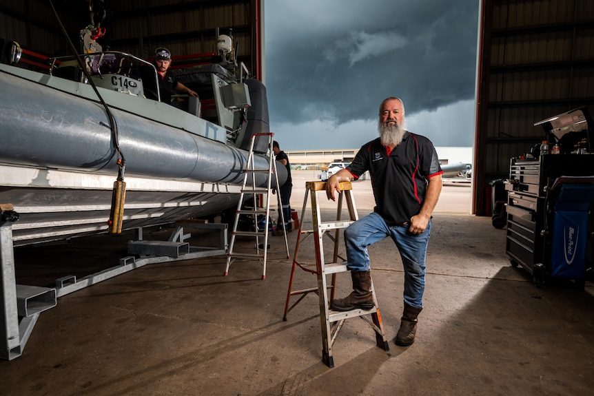 A man with a long grey beard stands next to a boat in a warehouse shed.