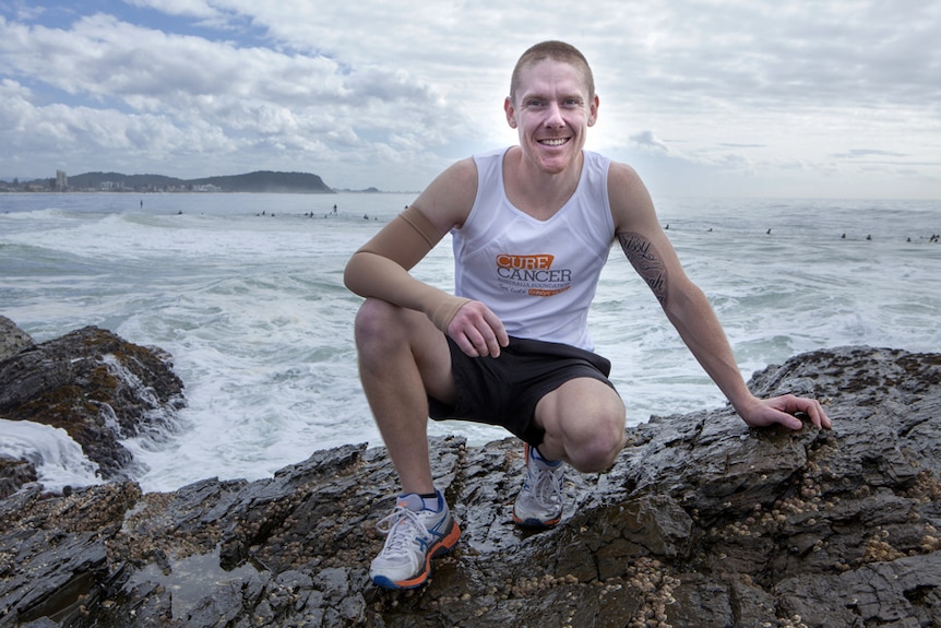 A man in running shorts and a singlet crouching on rocks in front of the ocean.