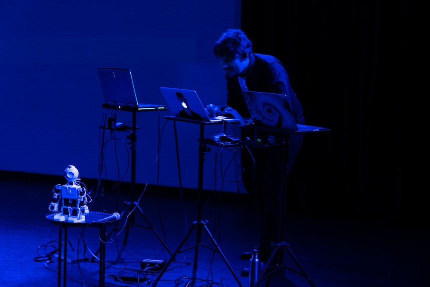 A man stands and operates a small robot using one of three laptops, which are all on stands