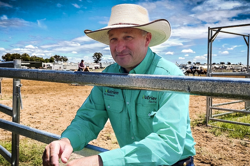 A man in cowboy attire leaning on a stockyard gate.