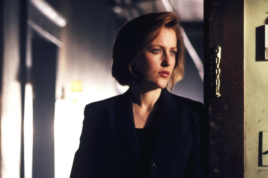 Dana Scully leans on a doorframe staring to the side of the camera.
