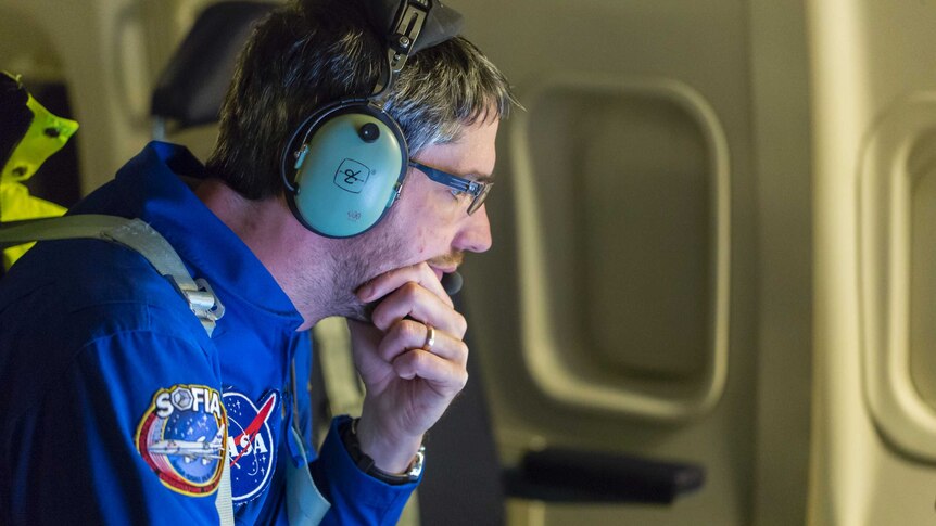 A man wearing a NASA-badged jumpsuit and headset looks intently at a device out of shot.