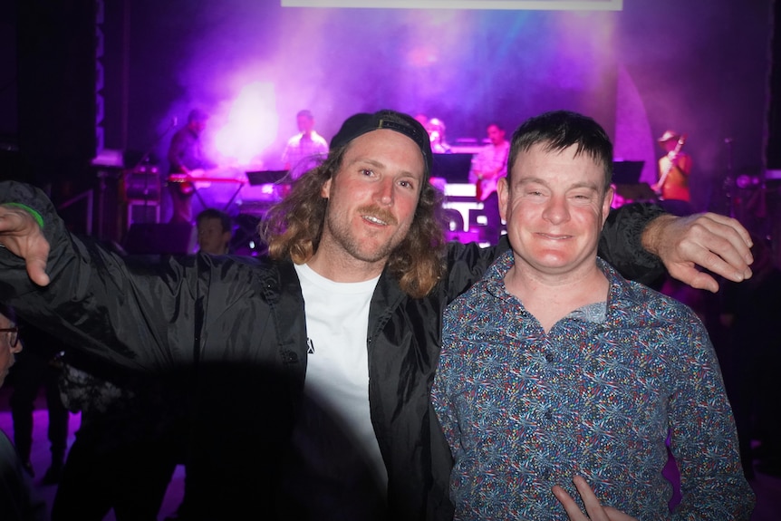 Two men stand in a nightclub smiling.