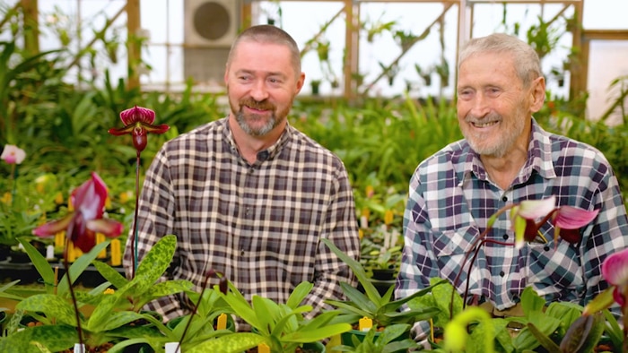 Two men smiling sitting in a greenhouse filled with orchids