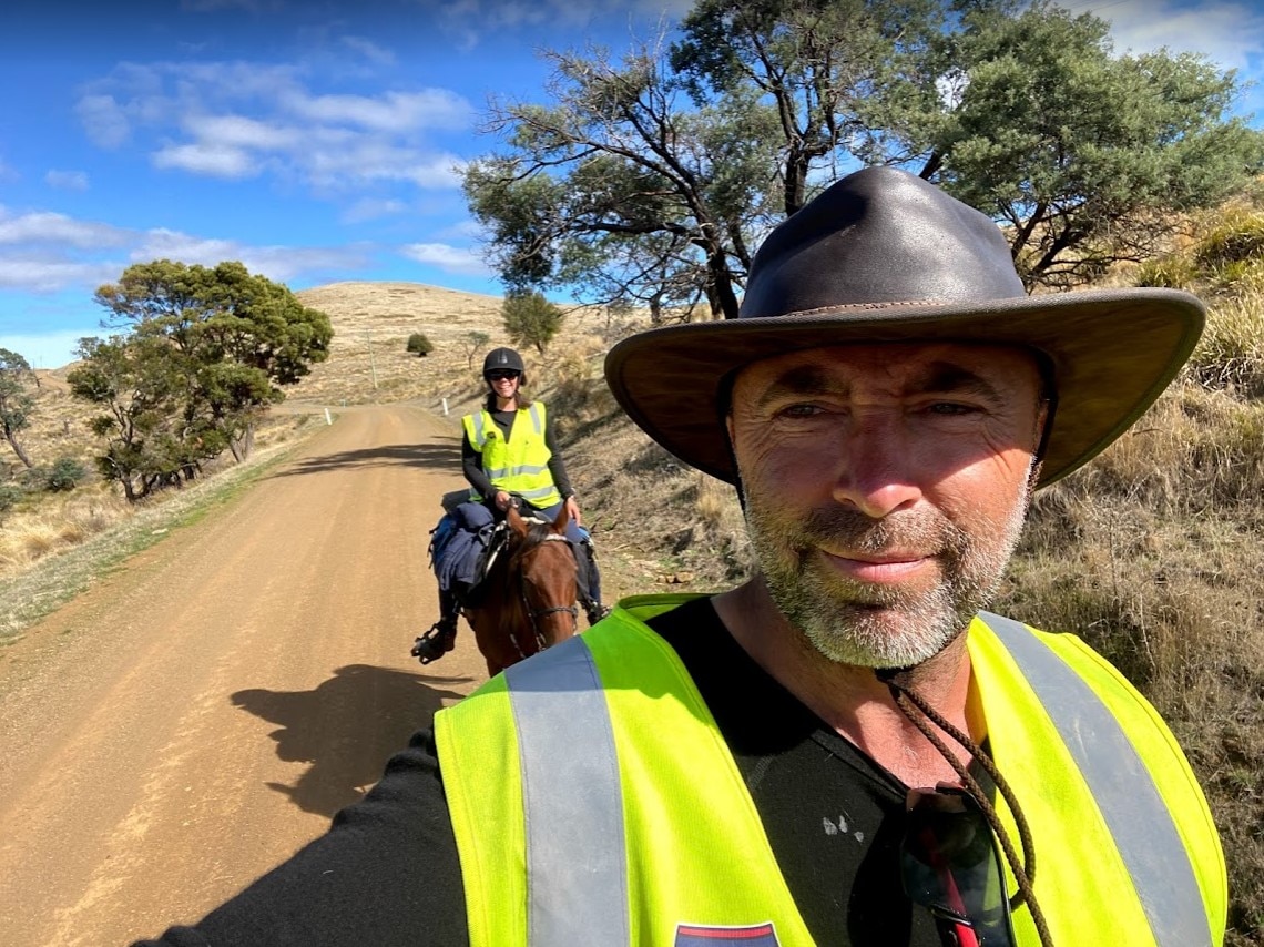 A man in an akubra type hat and high vis vest in the foreground, taking a selfie, a woman on horseback behind, smiling.