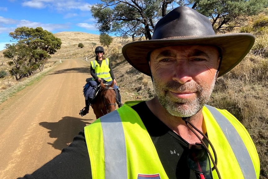 A man in an akubra type hat and high vis vest in the foreground, taking a selfie, a woman on horseback behind, smiling.