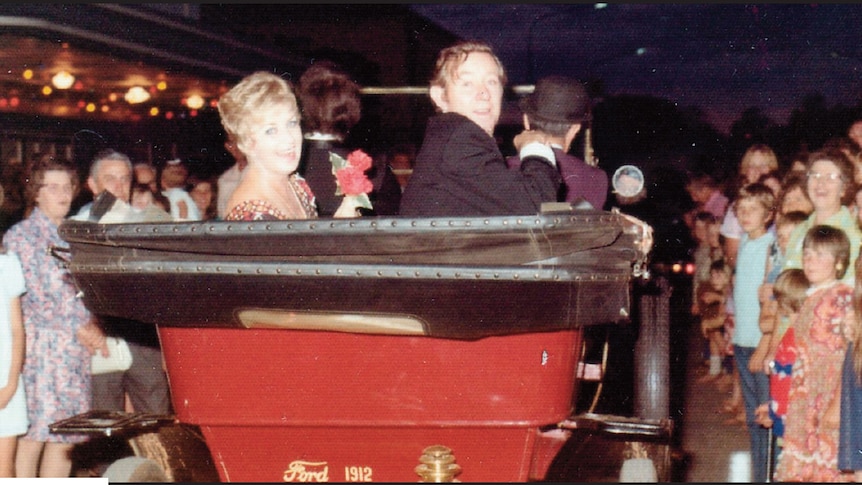 A man and woman sit in a vintage car with crowds looking on.