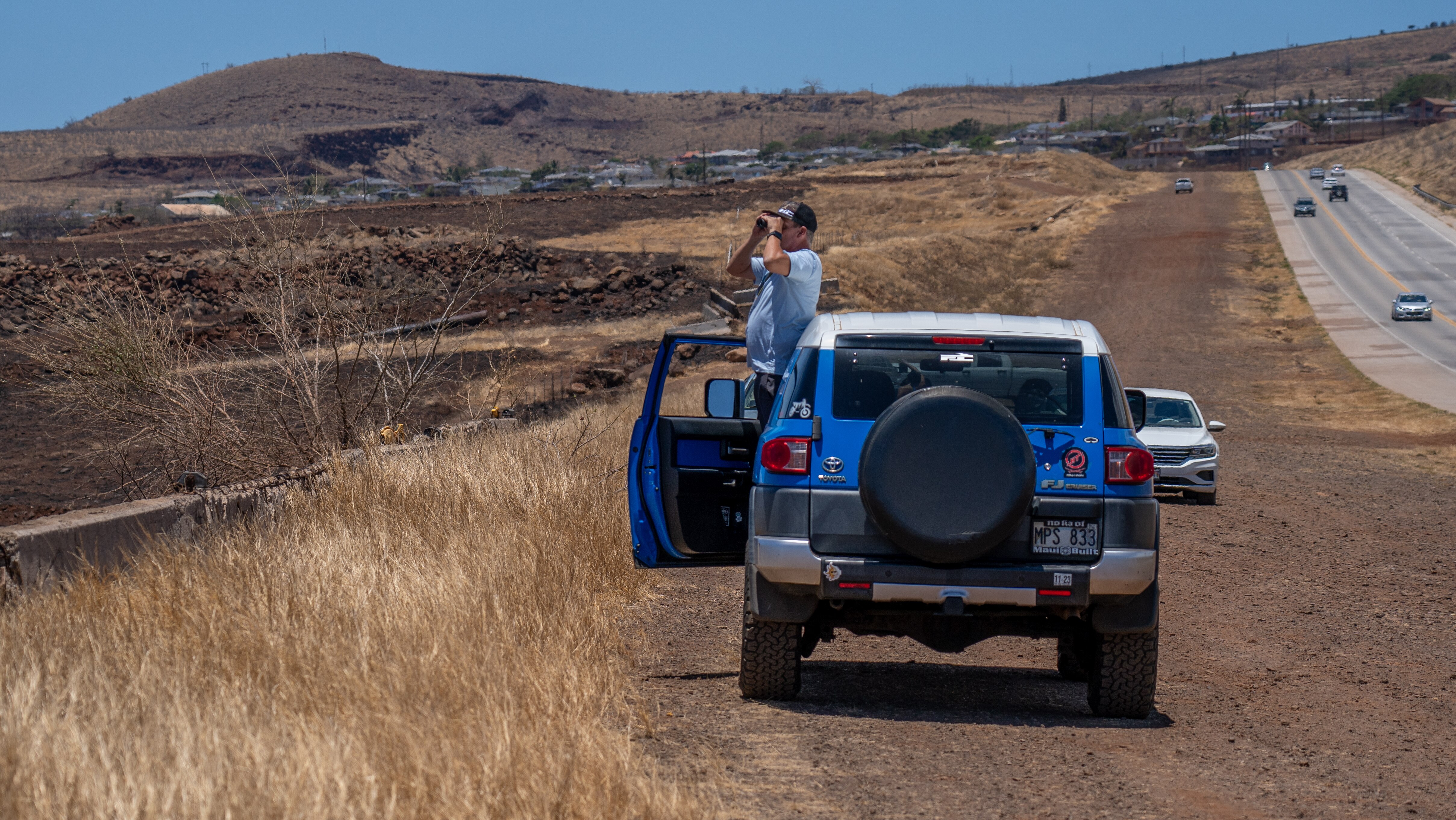 A man stands outside the door of his car and holds up binoculars. A burned town is in the distance.
