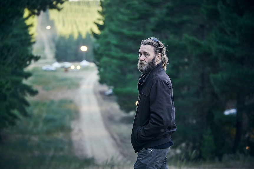 Middle-aged man white man with rustic beard, and long hair wearing black bomber jacket standing on a dirt road in bushy area.