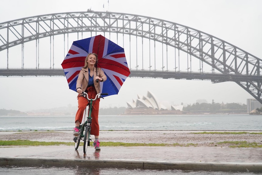Liz Truss sits on a bike while holding a Union Jack umbrella in front of the Sydney Harbour Bridge.