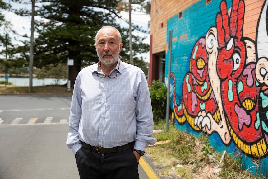 An elderly man stands by a painted wall.