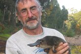 A man, wearing glasses, holds a platypus on the bank of a river